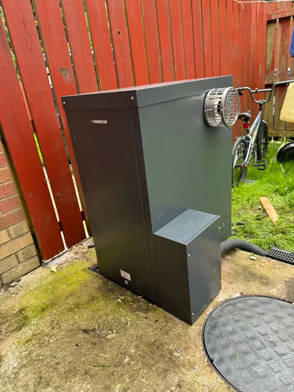Outdoor Warmflow oil boiler beside a red timber fence, flue terminal on the side
